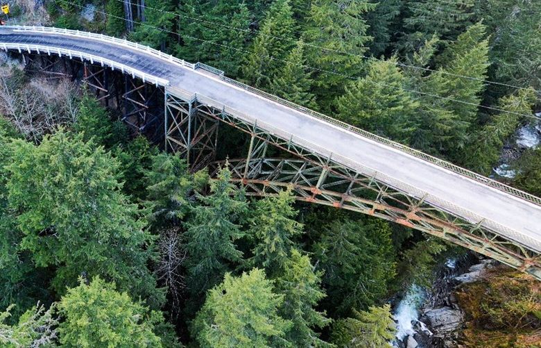 The SR 165 Carbon River Fairfax Bridge is a steel truss bridge supported by a lattice work of steel beams and columns. In this recent aerial image, rusting areas on the steel truss portions of the bridge are visible underneath the bridge deck.