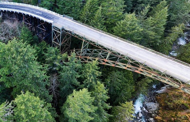 The SR 165 Carbon River Fairfax Bridge is a steel truss bridge supported by a lattice work of steel beams and columns. In this recent aerial image, rusting areas on the steel truss portions of the bridge are visible underneath the bridge deck.