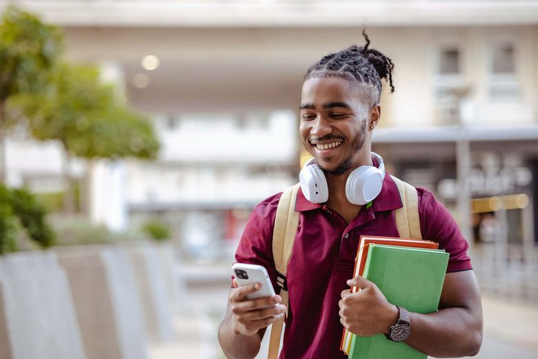 A young Black man with white headphones resting around his neck looks at his smartphone.