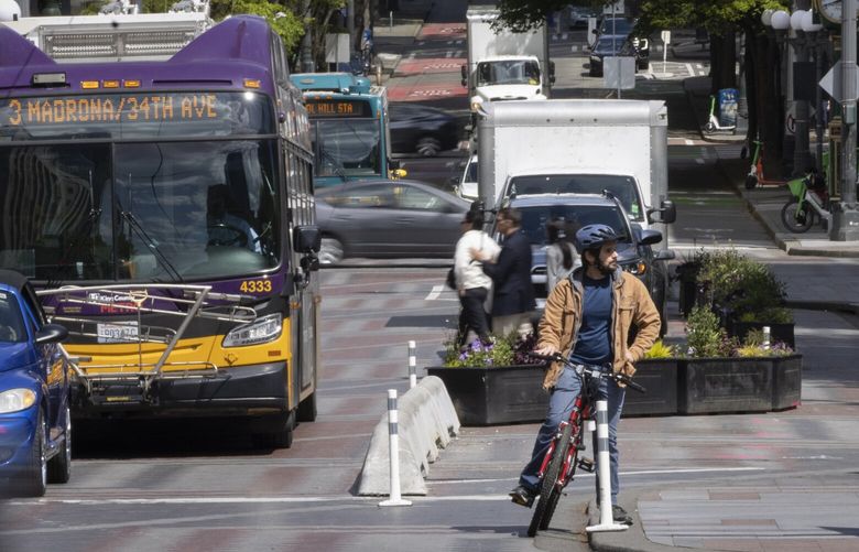 As traffic and pedestrians whiz by, a bicyclist waits in the Pine Street bike lane, where it meets the Fourth Avenue Bikeway, near  new concrete barriers at left, adjacent to Westlake Park (at right, out of photo), Friday, May 2, 20i25..

 After two vehicles hit bicyclists, the city quickly installed concrete barriers to better protect people on bikes. 230016
