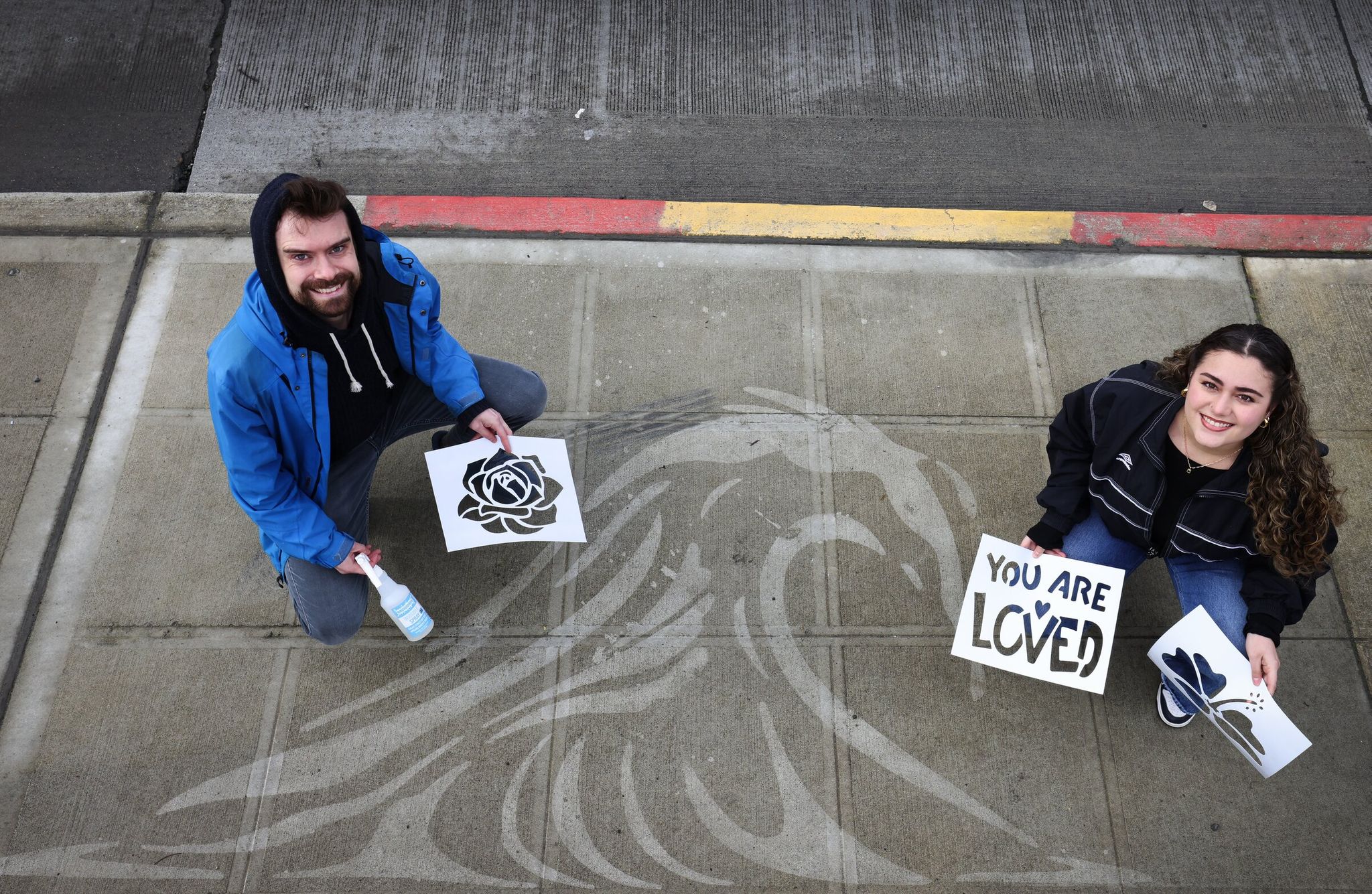 Rainworks founder Peregrine Church holding the ‘invisible spray’ and a stencil, and creative director Camille Kaputis, beside one of their sidewalk works on Monday at Beacon Hill. The tidal-wave installation appears in the rain. (Ken Lambert / The Seattle Times)