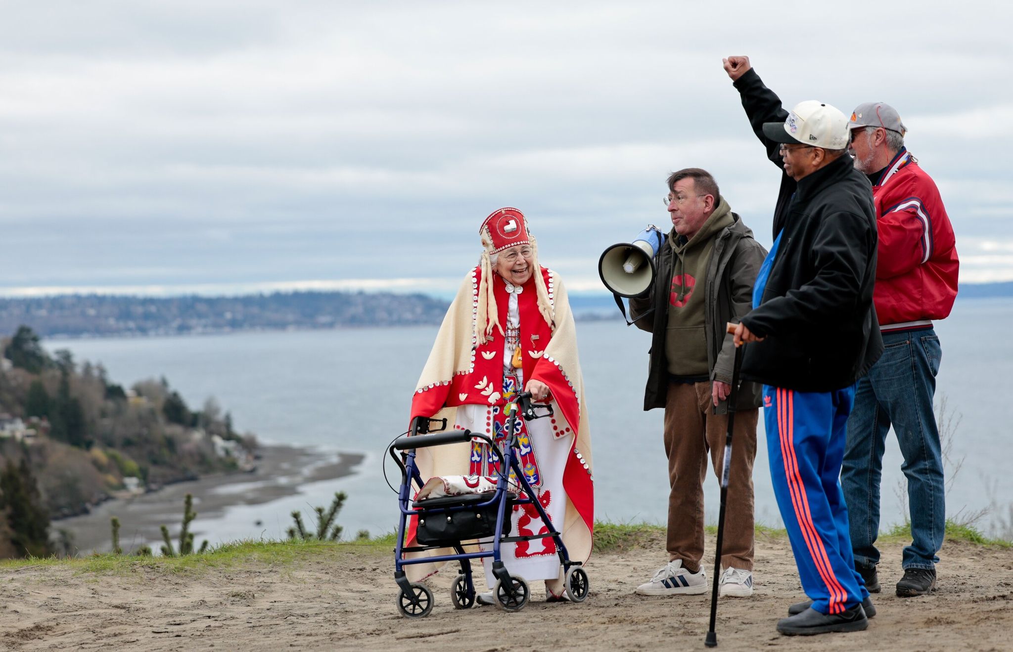 From left, Anna Haala (Tlingit); José Montaño, curator at Daybreak Star’s Sacred Circle Gallery; Tony Monroe, facility director for United Indians of All Tribes Foundation and Yakima tribal member; and Larry Gossett with his fist raised in the air, address the crowd during an opening ceremony at the sand dunes over looking the bluffs at Discovery Park in Seattle on Saturday. The United Indians of All Tribes Foundation commemorated the 1970 takeover of Fort Lawton, with a tribute to Larry Gossett and others who were activists from the 1970 occupation. (Karen Ducey / The Seattle Times)