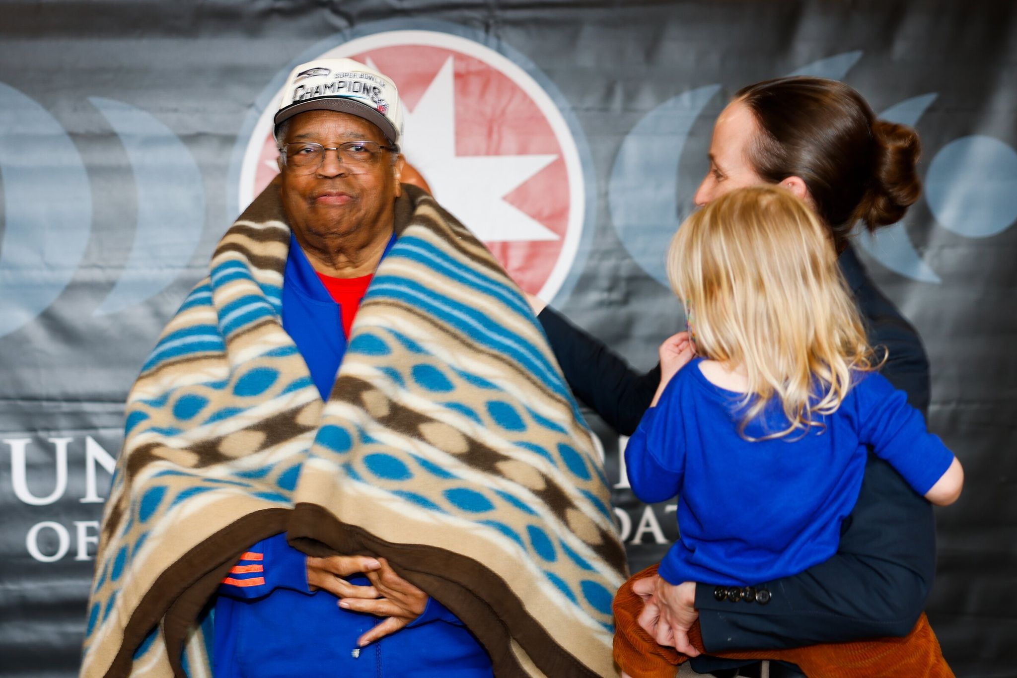 Larry Gossett, the last surviving member of the “Gang of Four,” which amplified minority representation in Seattle during the civil rights era, and a former King County Council member, is wrapped in a blanket by Seattle Mayor Kate Wilson, who is holding her daughter Josephine, during an event by the United Indians of All Tribes Foundation honoring the 1970 Fort Lawton takeover at the Daybreak Star Indian Cultural Center in Seattle on Saturday. (Karen Ducey / The Seattle Times)