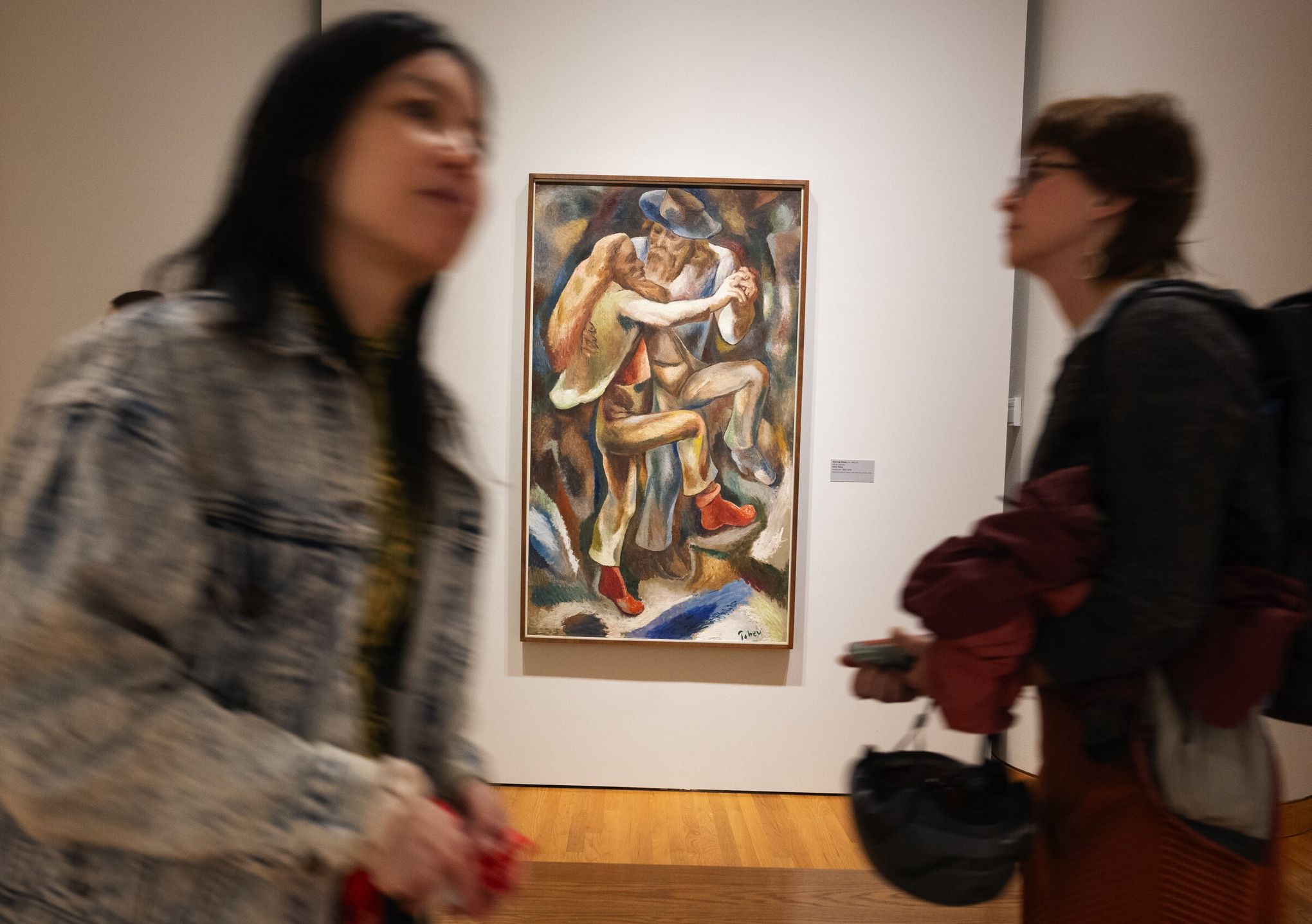 Two people walking past a framed painting of two dancers on a museum wall; both are slightly blurred, while the artwork is in focus. The gallery features wood flooring and neutral walls.