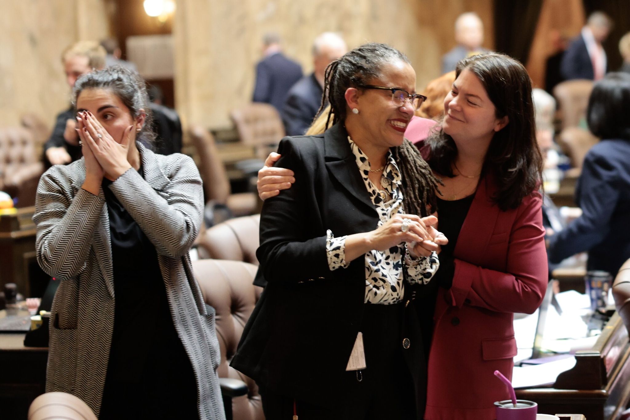 Эйприл Берг (в центре) обнимает представительница Лиз Берри (справа), в то время как представительница Дарья Фаривар (слева) тоже празднует после дебатов Палаты представителей по «налогу на миллионеров», принятых в здании Капитолия штата в Олимпии во вторник, 10 марта 2026 года. (Карен Дьюси / The Seattle Times)