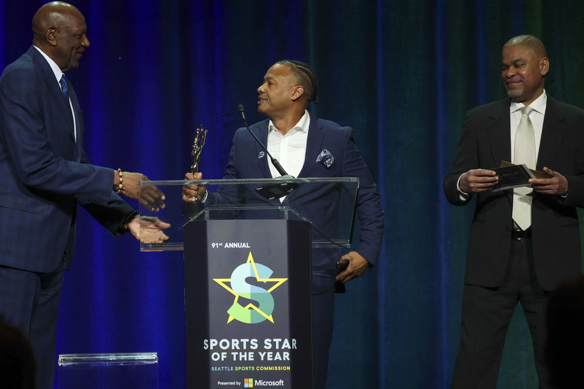NBA star Spencer Haywood (left) presents the Male Athlete of the Year award to Seahawks community and legends relations vice president Mario Bailey (center), who accepted the award on behalf of Jaxon Smith-Njigba, while former NBA player Eldridge Rekasner (right) looks on during the 91st annual Seattle Sports Star of the Year Awards at the Seattle Convention Center Summit in downtown Seattle on Friday, March 13, 2026. (Ivy Ceballo / The Seattle Times)