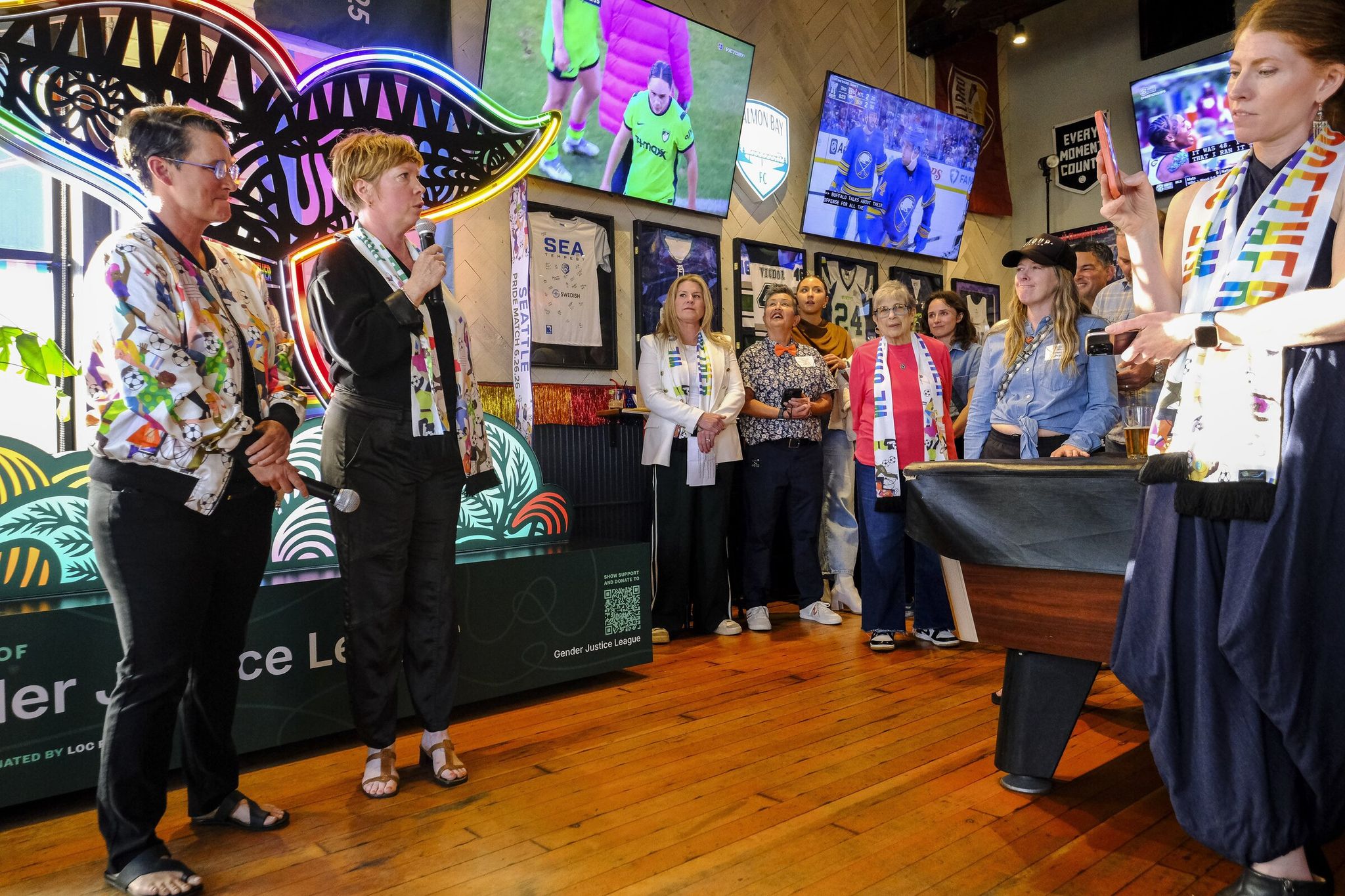 Jen Barnes, owner of Rough & Tumble Pub, and Hedda McLendon, senior vice president of legacy for the local organizing committee, address the crowd at the popular Ballard bar before the debut of videos celebrating Pride. (Dean Rutz / The Seattle Times)