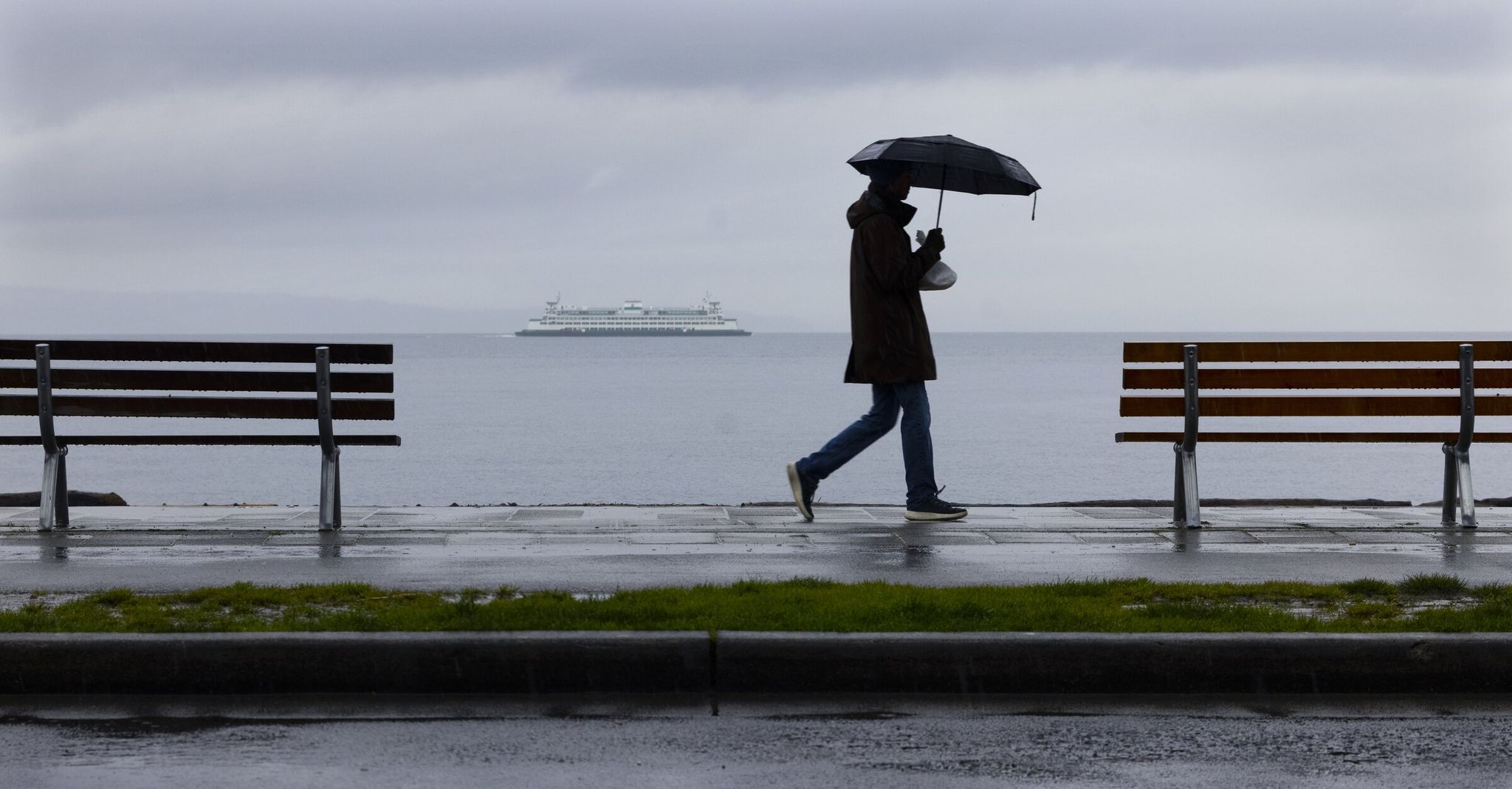 As a Washington State Ferry heads away from Seattle, a  person walks on Alki Avenue Southwest, on a “gray and drippy” day earlier this year. (Ellen M. Banner / The Seattle Times)