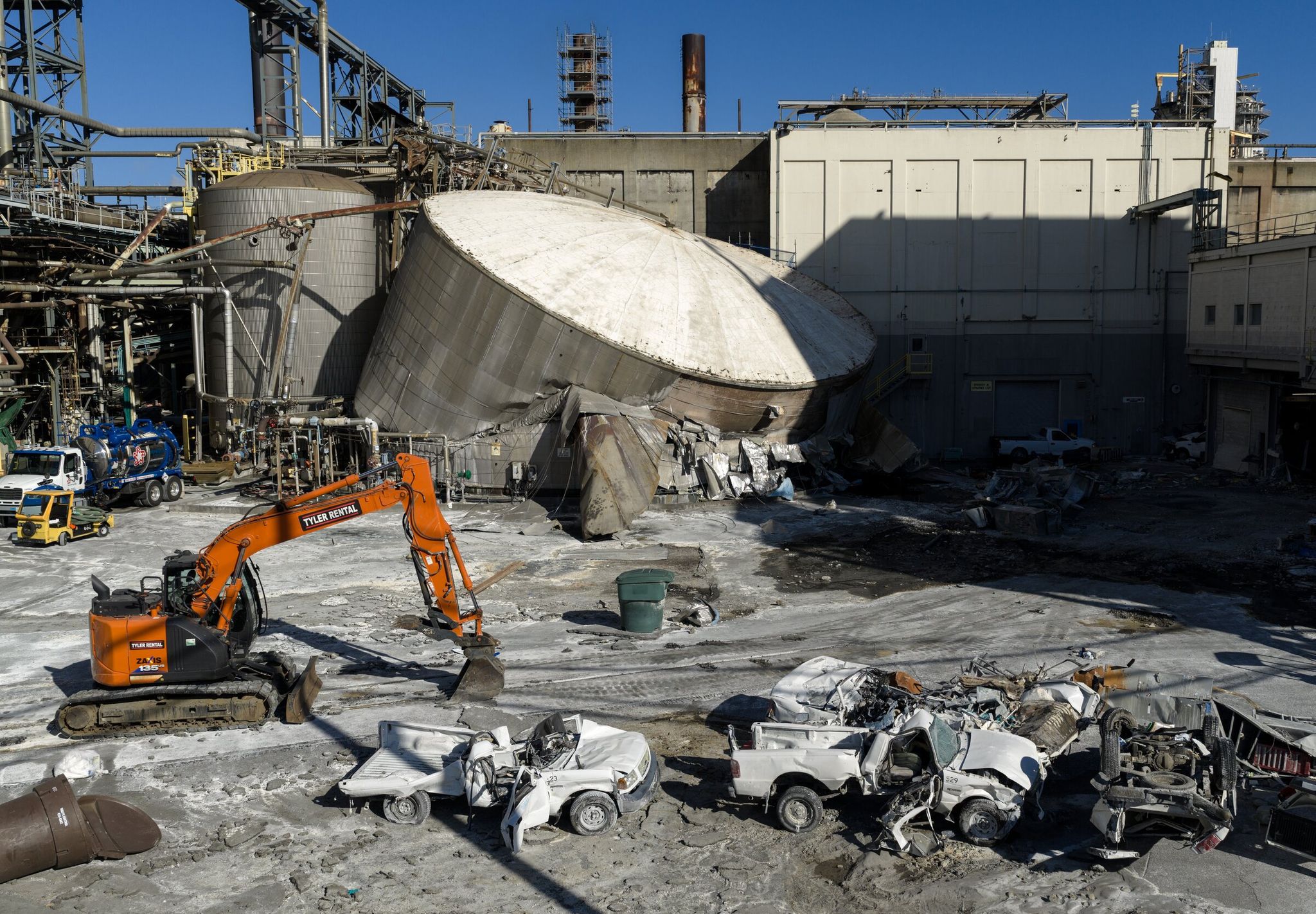 An excavator stands beside a pile of mangled pickup trucks and a collapsed chemical tank at the Nippon Dynawave Packaging Co. site on Sunday, May 31, 2026, in Longview. The implosion on Tuesday caused a release of a highly caustic chemical that killed 11 workers at the pulp and paper plant. (Nick Wagner / The Seattle Times)