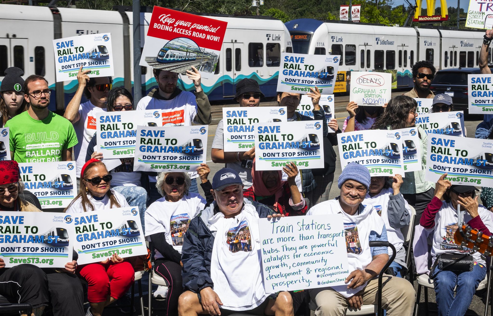 A light rail 1 Line train speeds by along Martin Luther King Jr. Way South as people demanding more stations gather at an event hosted by Southend Transit Justice Coalition in May. (Ken Lambert / The Seattle Times)