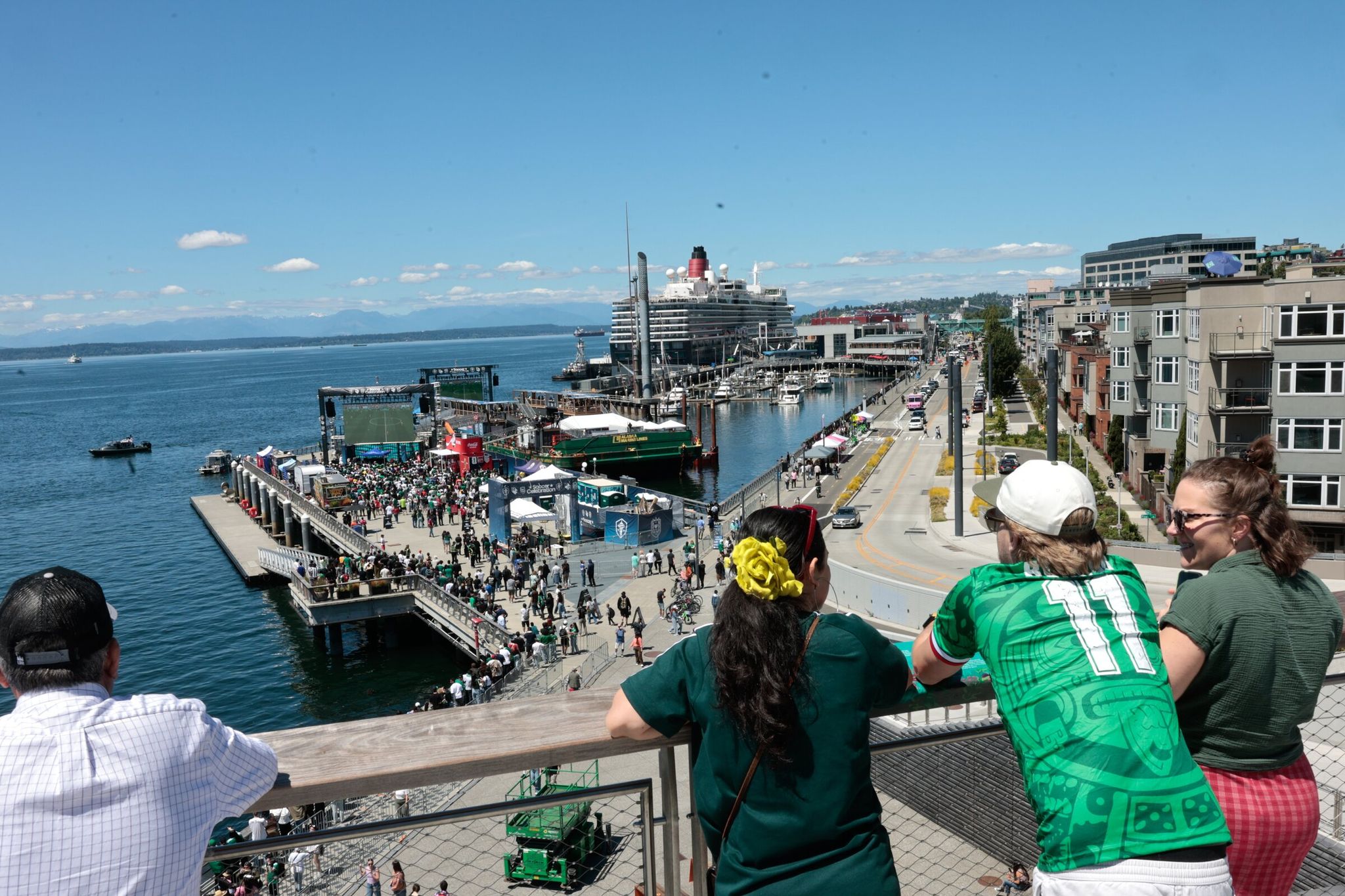 Taking in views of the Olympic Mountains, the World Cup plays on a big screen and the FIFA floating barge is visible: Jessica Hernandez (left), and John Elliano and Maya Elliano. They are pictured overlooking Pier 62 in Seattle on June 10. (Karen Ducey / The Seattle Times)