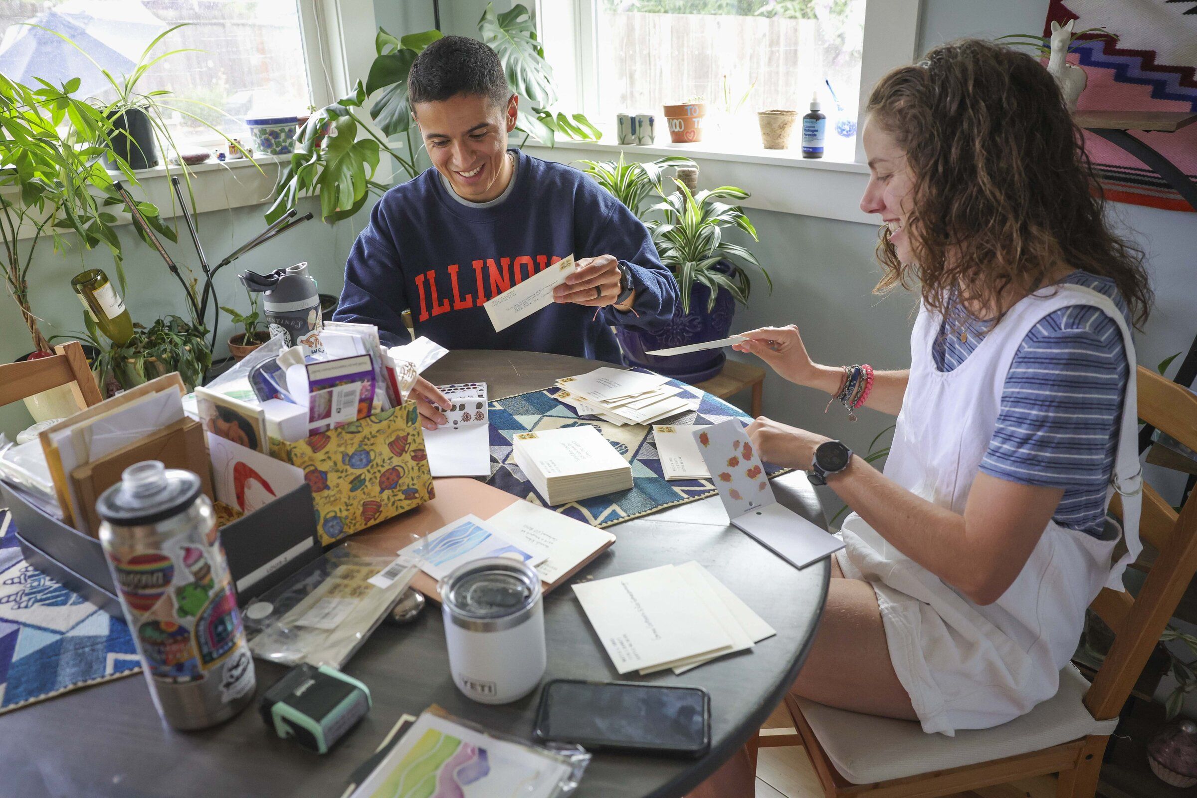 Jorge Aranda, left, and Mattie Gottbrath, right, stuff envelopes with save the date cards for their wedding at their Seattle home on Saturday, June 27, 2026. (Ivy Ceballo / The Seattle Times)