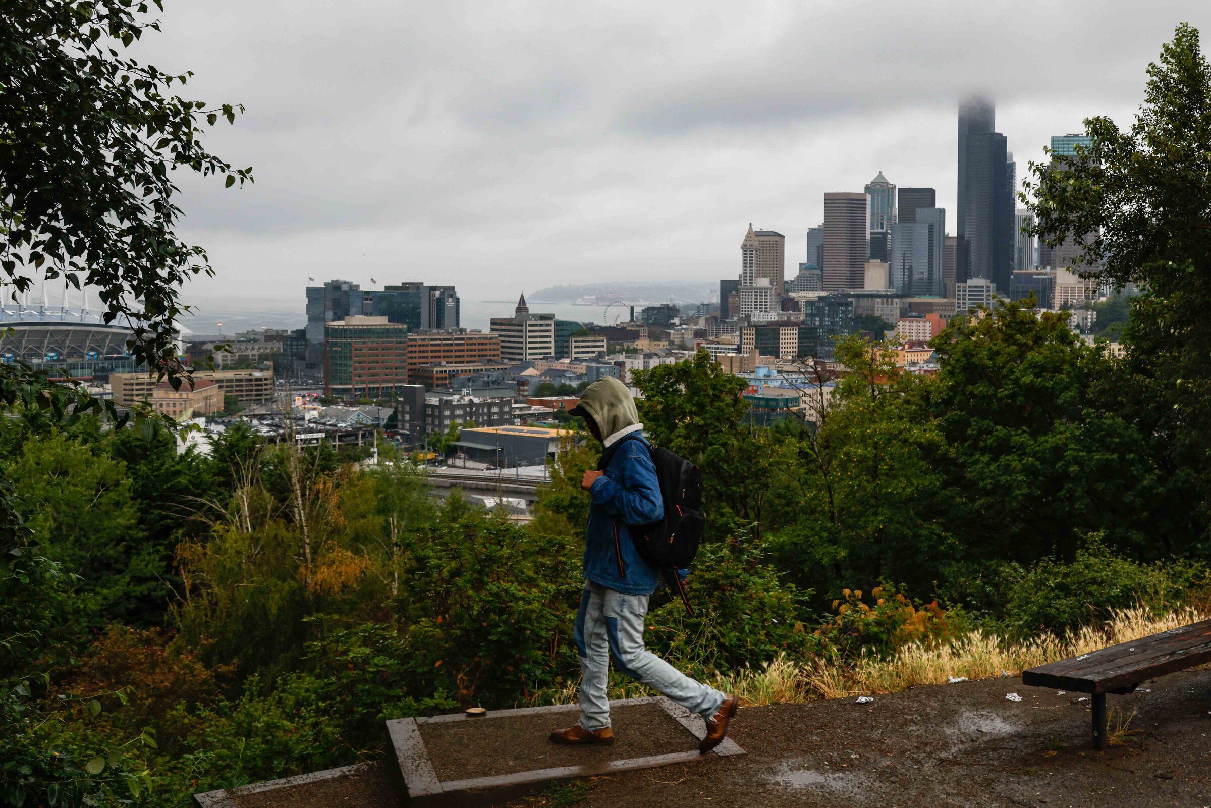 A pedestrian walks through Jose Rizal Park with a view of a cloudy Seattle during a storm on Thursday. (Andrew Burke-Stevenson / The Seattle Times)