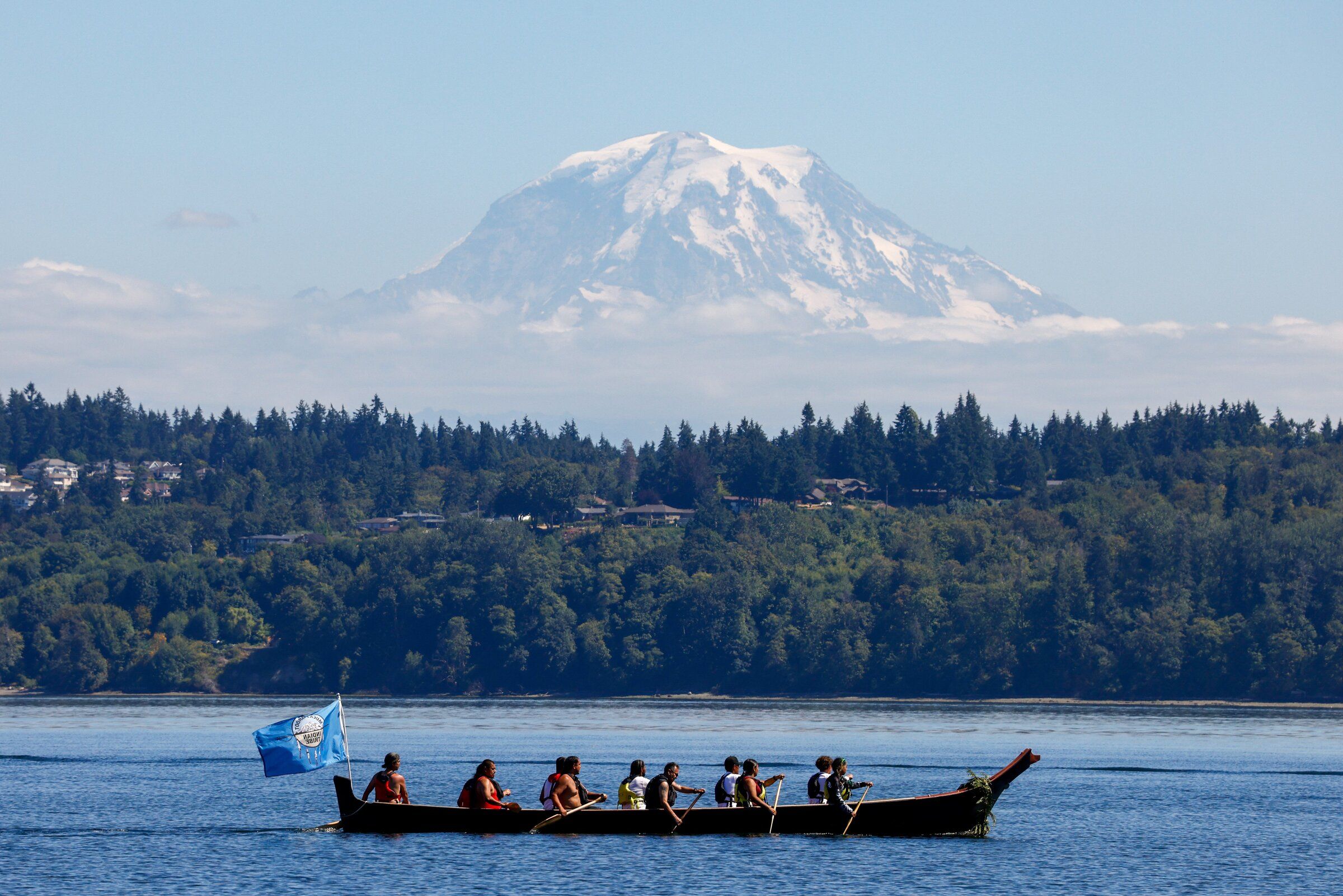 The Muckleshoot Canoe Family passes təqʷubəʔ (Mount Rainier) in yəx̌ʷuləʔ səli (Eagle Spirit) as they pull from Alki Beach to Puyallup through the Salish Sea on July 30. The canoe gained its name after an eagle swooped down in front of it on a previous journey, according to Canoe Family manager Autumn MaGee. (Andrew Burke-Stevenson / The Seattle Times)